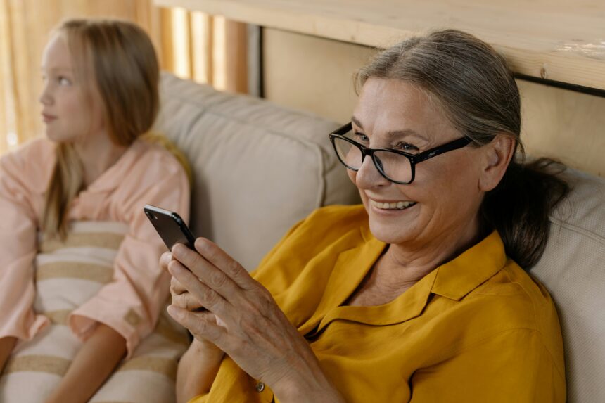 A smiling elderly woman with glasses holds a smartphone while sitting on a couch next to a young girl, potentially living with autism spectrum disorder, in a cozy, relaxed environment.