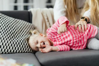 A young child laughing while being tickled by an adult on a sofa, wearing a pink and white striped shirt.