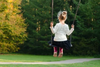 A young girl sitting on a swing, facing away, with a forest in the background.